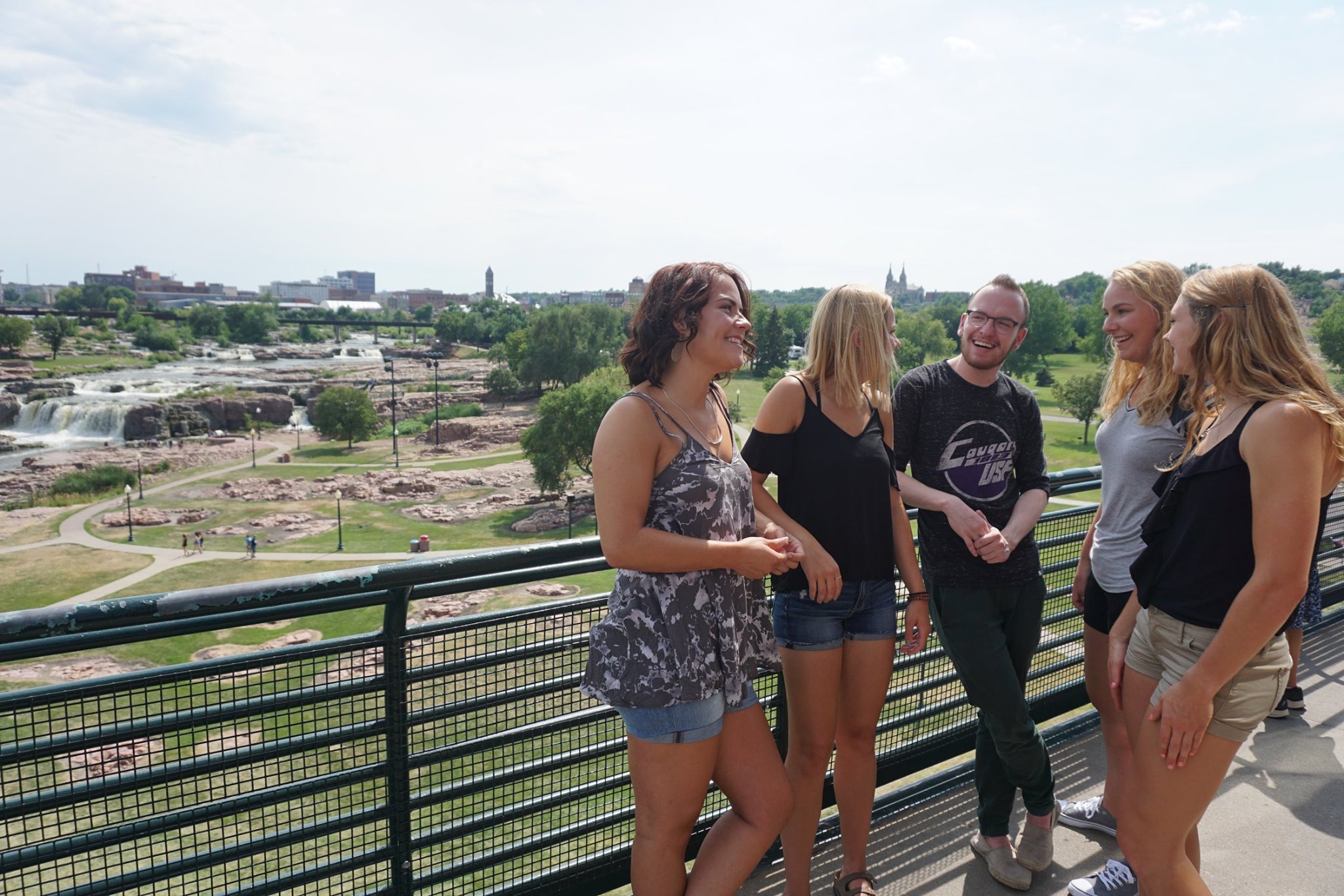 A group of five people stand on an outdoor walkway with a scenic view in the background. They are smiling and engaged in conversation. In the distance are waterfalls, greenery, and a city skyline under a partly cloudy sky.