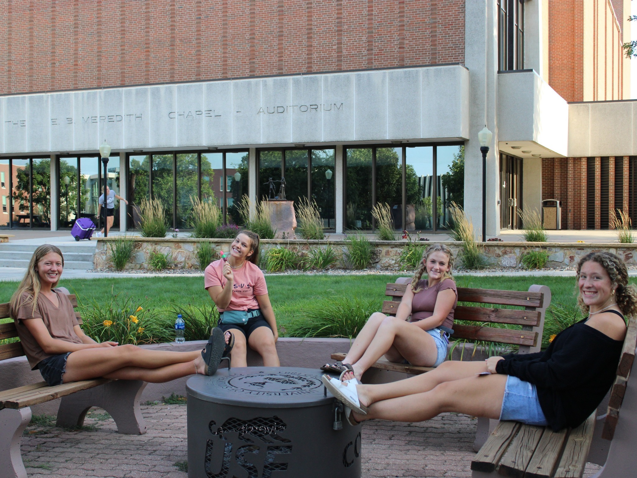 Four people are sitting on benches around a small fire pit, smiling and interacting. They have their feet propped up on the edge of the pit with relaxed postures. A modern building labeled "Meredith Chapel-Auditorium" is in the background, along with some greenery.