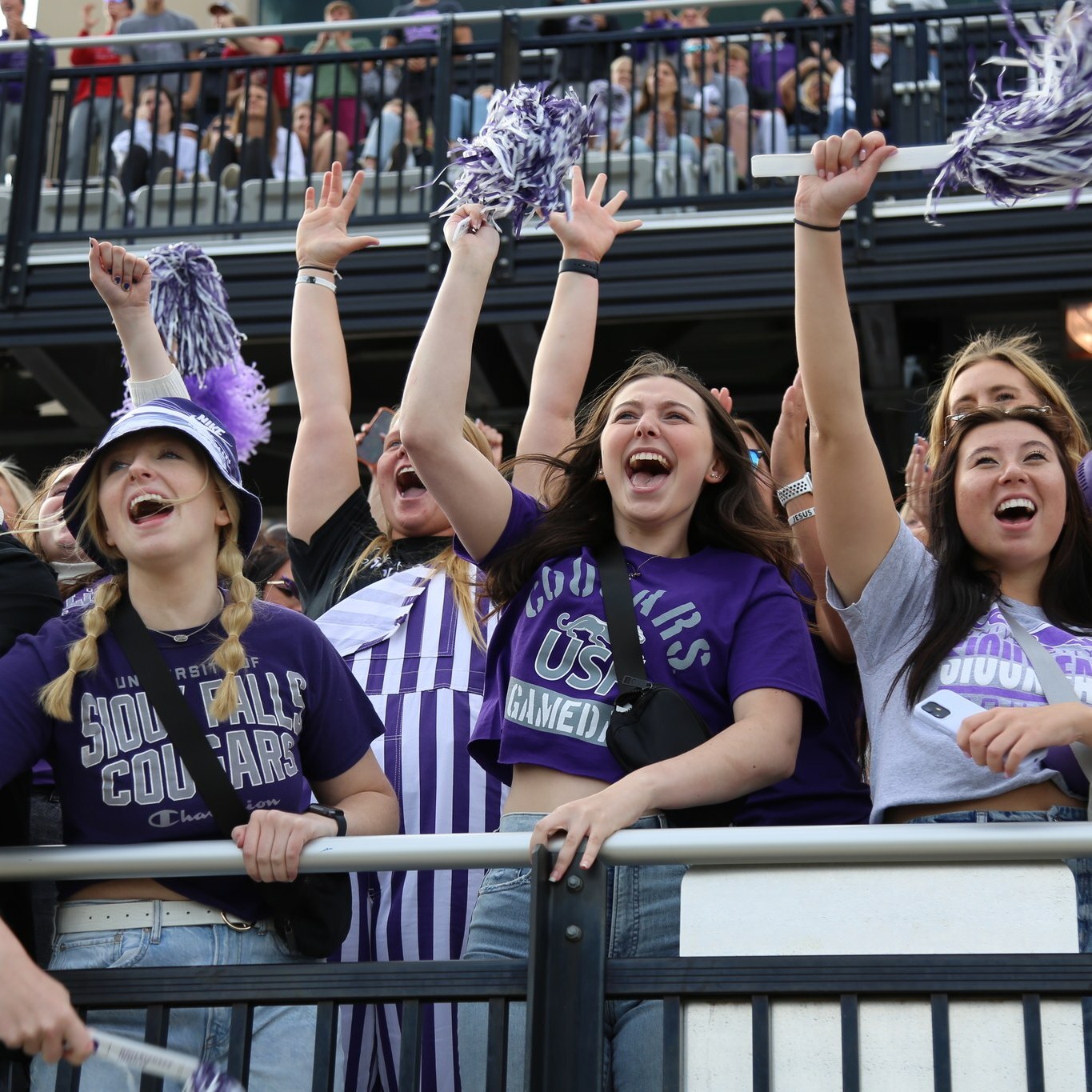 A group of enthusiastic fans wearing University of Sioux Falls clothing cheer and wave pompoms in the stands at a sports event. They are all smiling and appear to be extremely excited, with more fans visible in the background.