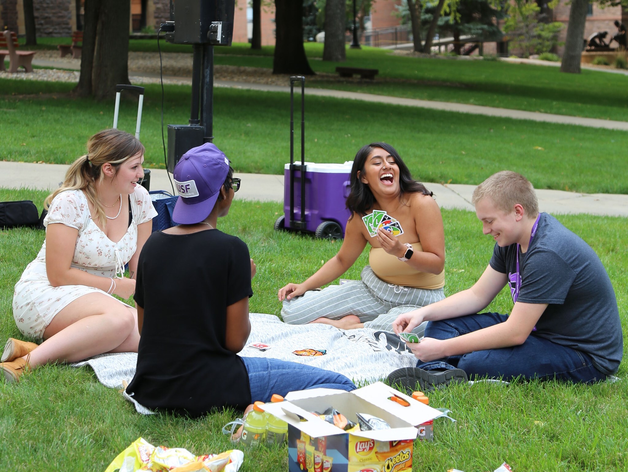 Four people are sitting on a picnic blanket on a grassy lawn, laughing and playing a card game. Snacks and drinks are spread out nearby. One person holds up several cards with a big smile, while the others join in the laughter. Trees and a building are in the background.