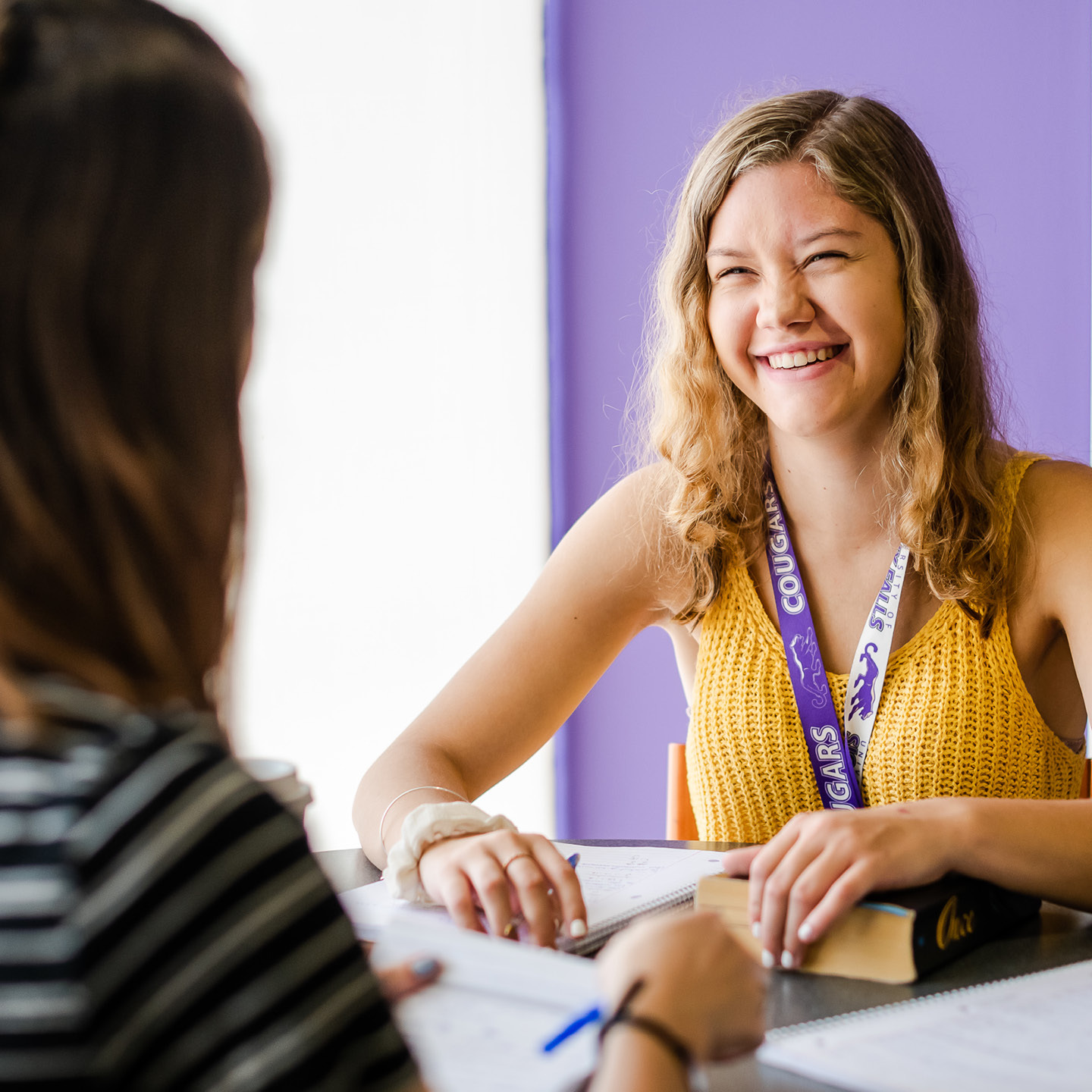 A young woman with long blonde hair, wearing a yellow sleeveless top and a "COUGARS" lanyard, smiles while sitting at a table with an open book in front of her. She faces another person who has their back to the camera. The background wall is purple.