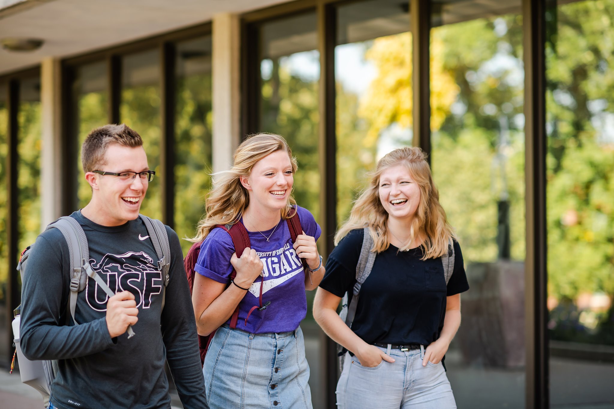 Three students are walking together and smiling outside a building with large windows. One student is wearing glasses and a dark shirt, the second student has a purple shirt and holds a phone, and the third student is dressed in black, all carrying backpacks.