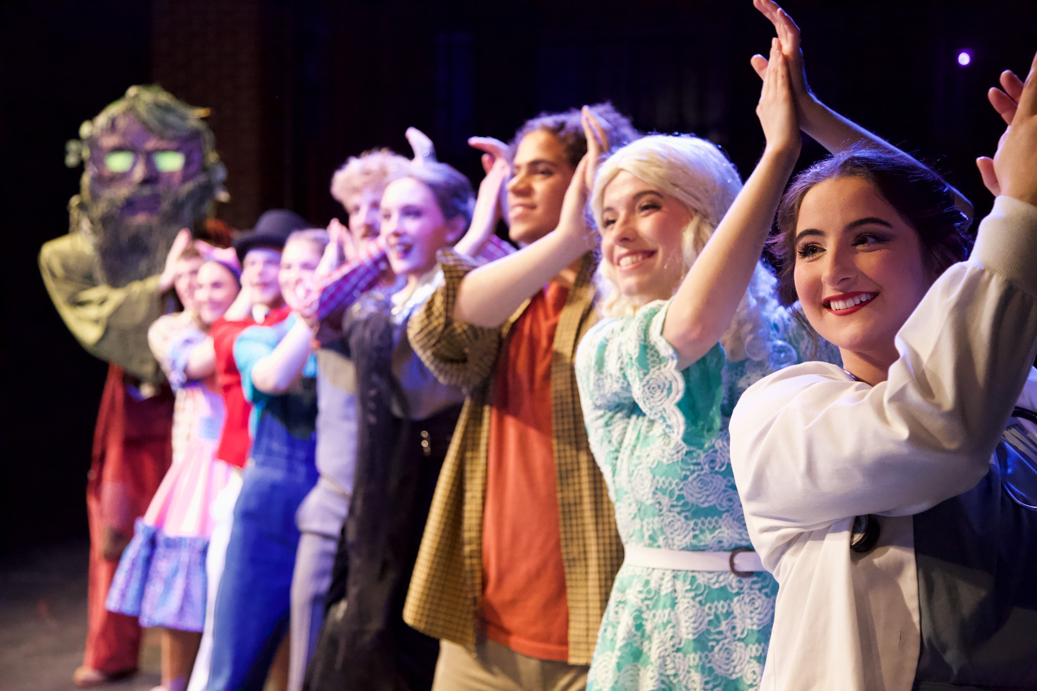 A group of performers dressed in colorful costumes stand in a line on stage, smiling and clapping their hands above their heads during a curtain call. The background is dimly lit with stage lights focused on the performers, highlighting their faces and attire.