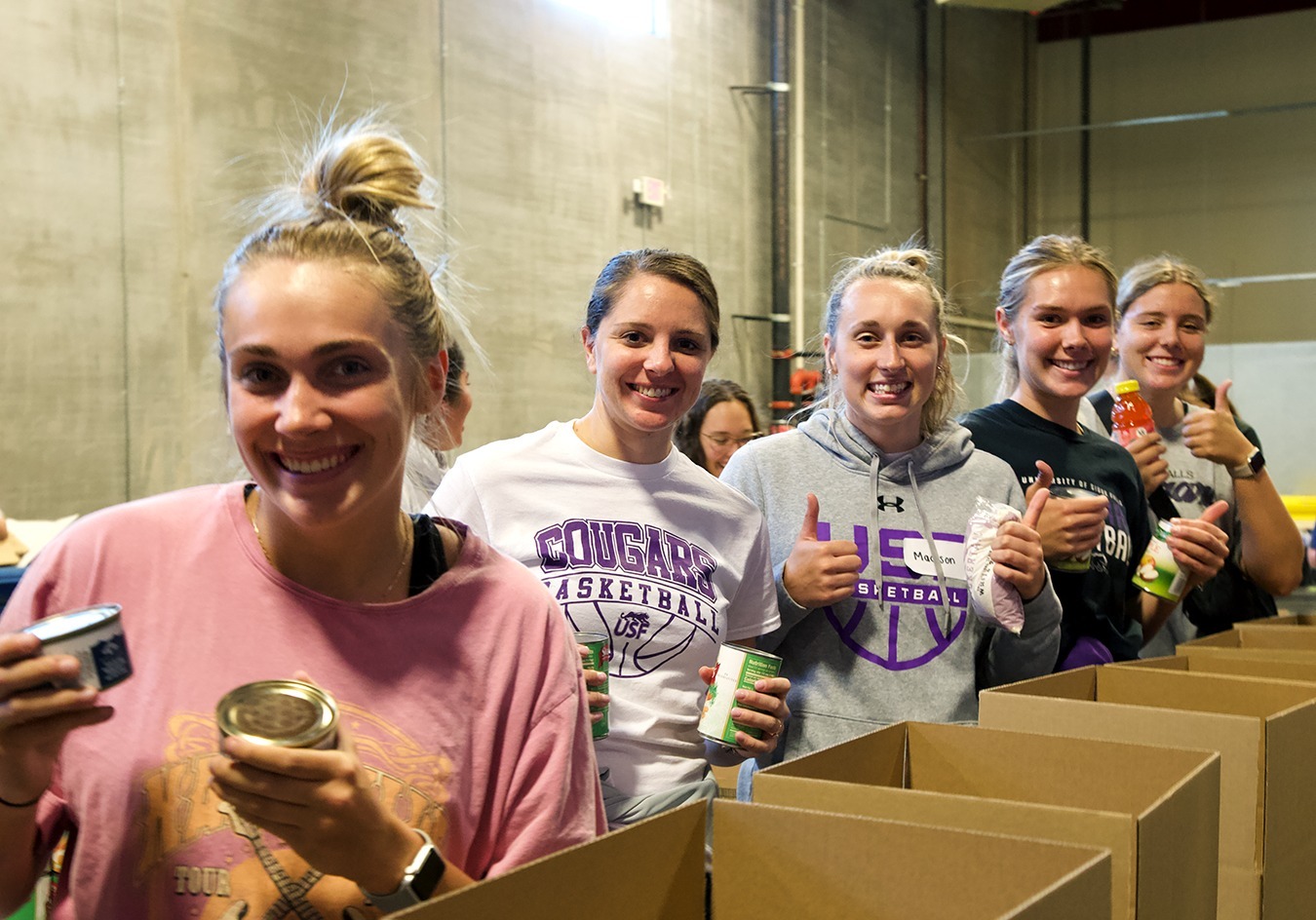 A group of five people, all women, stand in a warehouse, smiling and holding canned goods. Each person has their thumbs up, showing a positive and enthusiastic attitude. They are packing items into large cardboard boxes.