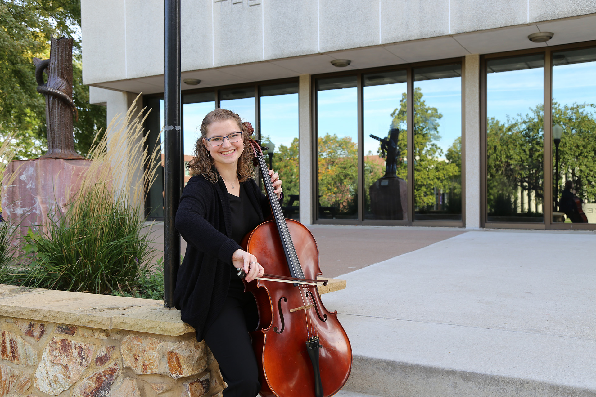 A woman with curly hair and glasses, dressed in black, smiles while playing a cello outside a building with large windows. She is seated on a stone ledge next to ornamental grass and a sculpture, enjoying the outdoor setting.