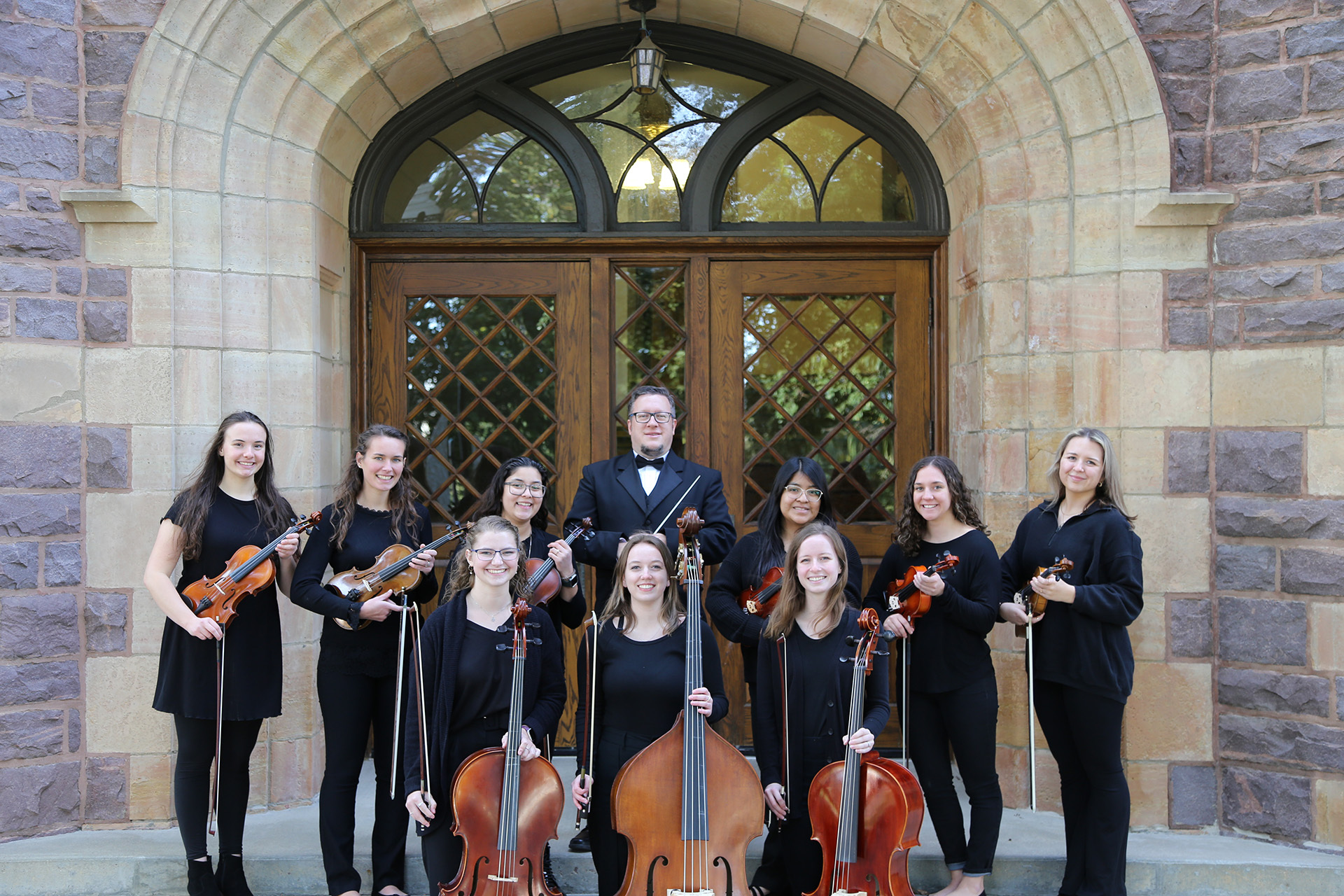 A group of nine musicians dressed in black attire, eight women and one man, stand in front of a large, arched wooden door. They are holding string instruments, including violins, cellos, and a viola. The building behind them has stone walls and a gothic architectural style.