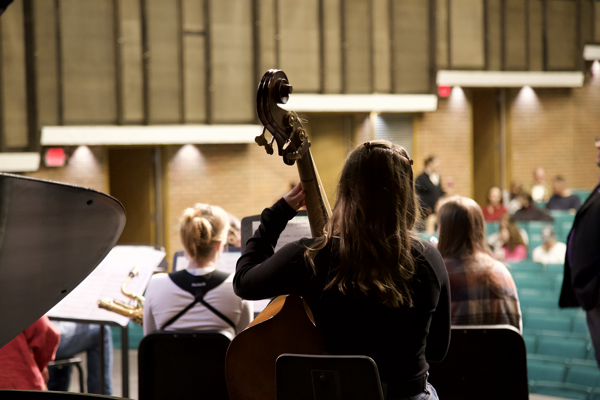 A group of young musicians is seated in a rehearsal space. A female musician in the foreground holds a double bass, her back facing the camera. Other musicians sit with assorted instruments, such as trumpets and cellos, and an audience area is visible in the background.