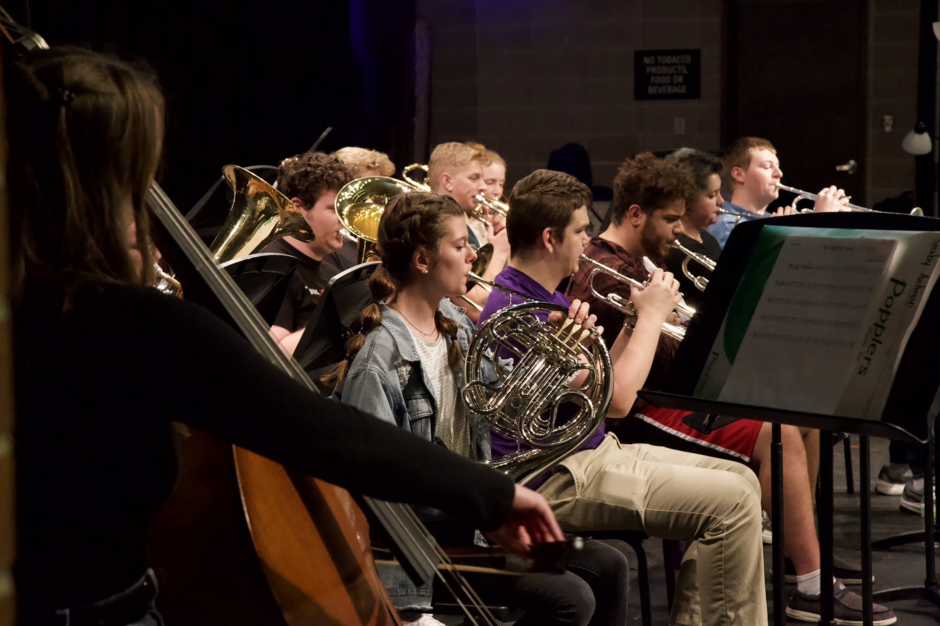A group of musicians is performing on stage, playing various brass and string instruments, including trumpets, trombones, French horns, and a double bass. The performers are seated and focused on their sheet music while playing. The background is dimly lit.