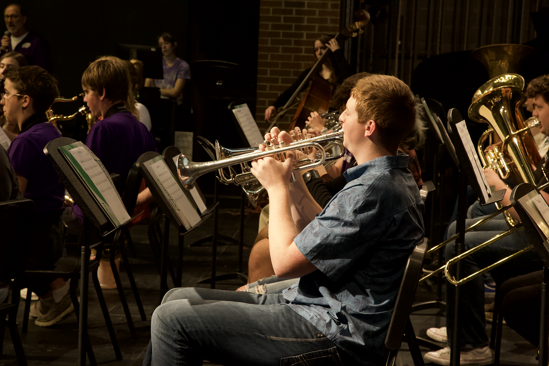 A young person plays a trumpet while seated in a musical ensemble. They are wearing a blue shirt and are surrounded by other musicians playing a variety of instruments, including trombones and a double bass. Sheet music is visible on stands.