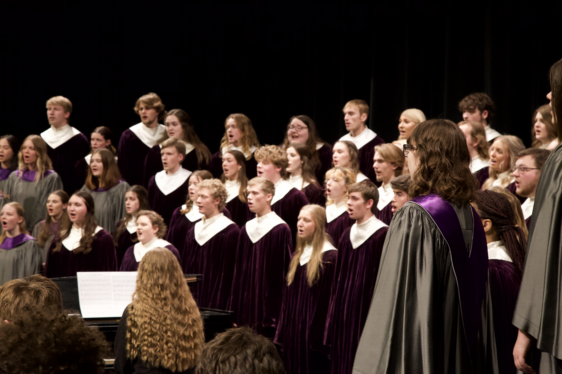 A large choir dressed in purple and gray robes performs on stage. Some members wear white collars. A person stands in the foreground wearing a gray robe with a purple sash. A pianist accompanies the choir, and an audience is seated in front.