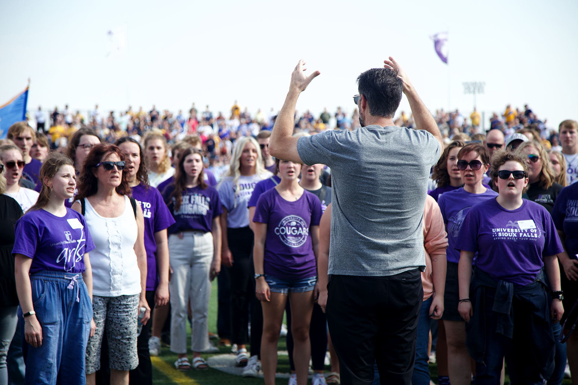 A crowd of people, many wearing purple shirts, stands outdoors on a sunny day. A person in a gray shirt, with their back facing the camera, appears to be addressing or leading the group. A large crowd is visible in the background, seated on bleachers.