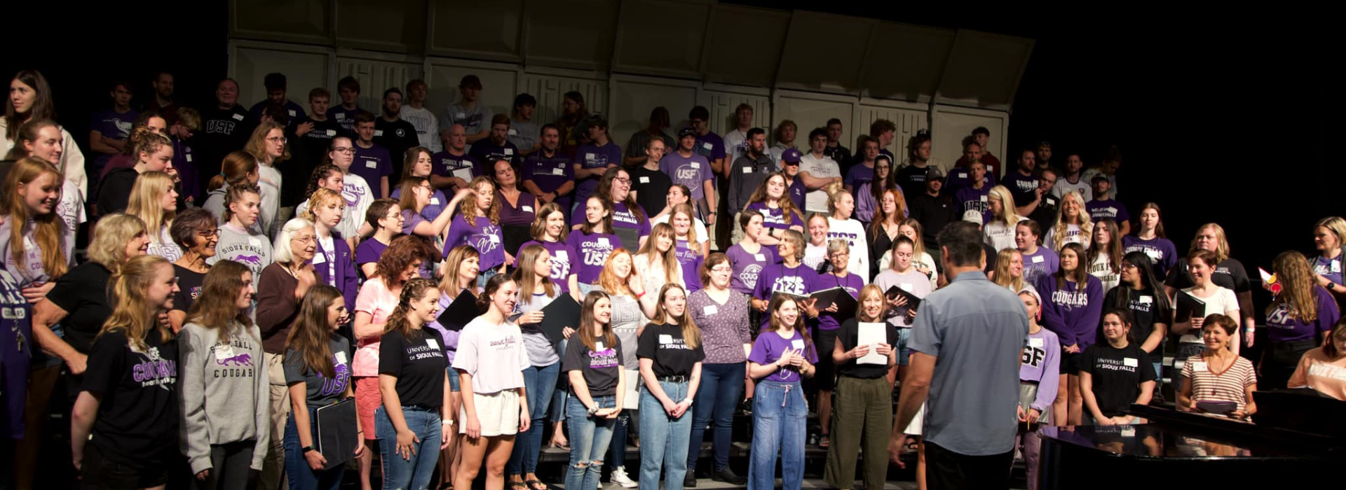 A large group of people, mostly wearing purple shirts with some in other colors, stand in several rows on multi-tiered stands. They are facing a man in a gray shirt and jeans who is standing with his back to the camera, appearing to address or conduct them.