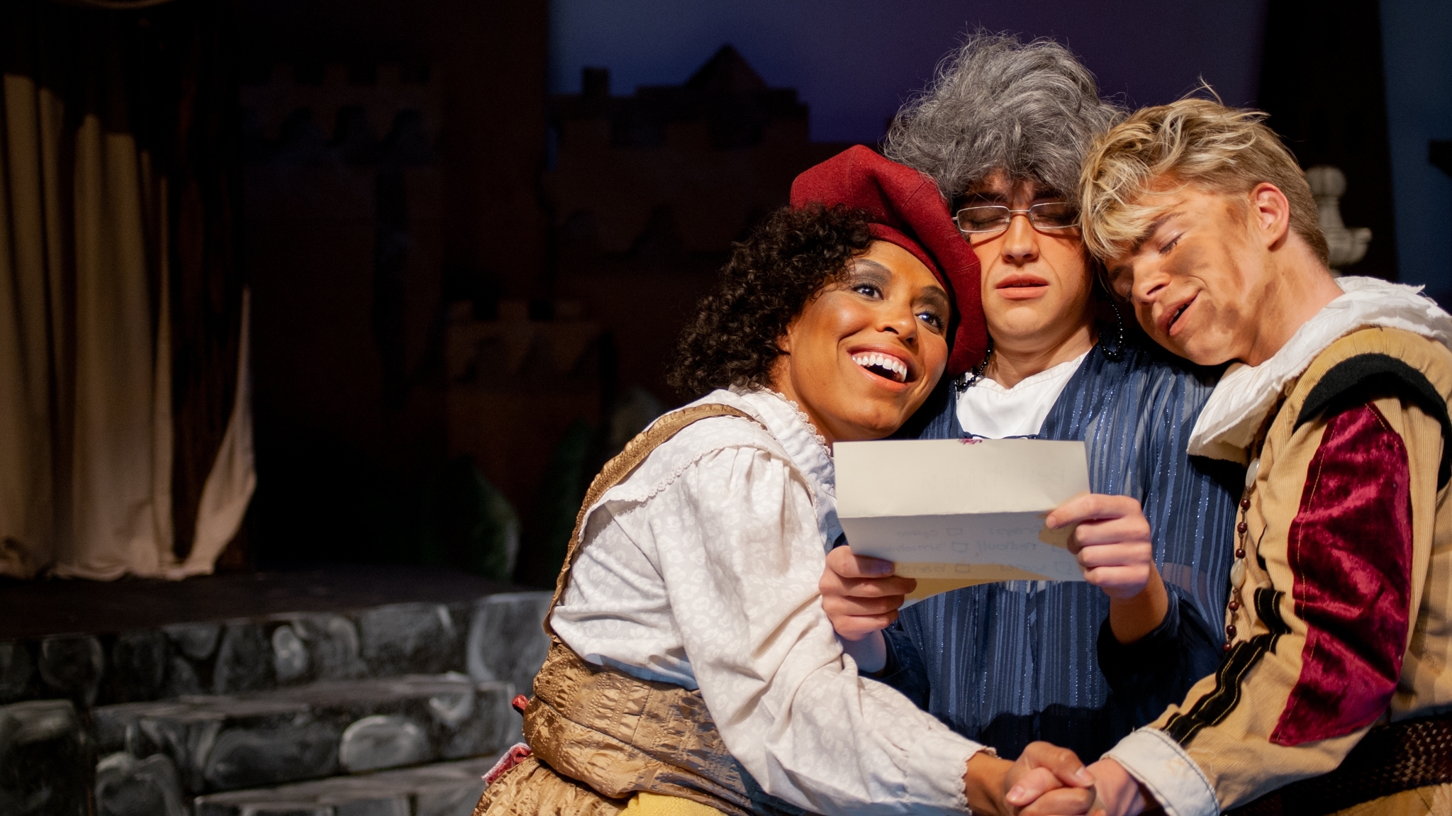 Three actors on stage, dressed in Renaissance-period costumes. The actor on the left holds a parchment while smiling. The actor in the middle, wearing a grey wig, frowns at the parchment. The actor on the right rests their head on the middle actor's shoulder, looking content.