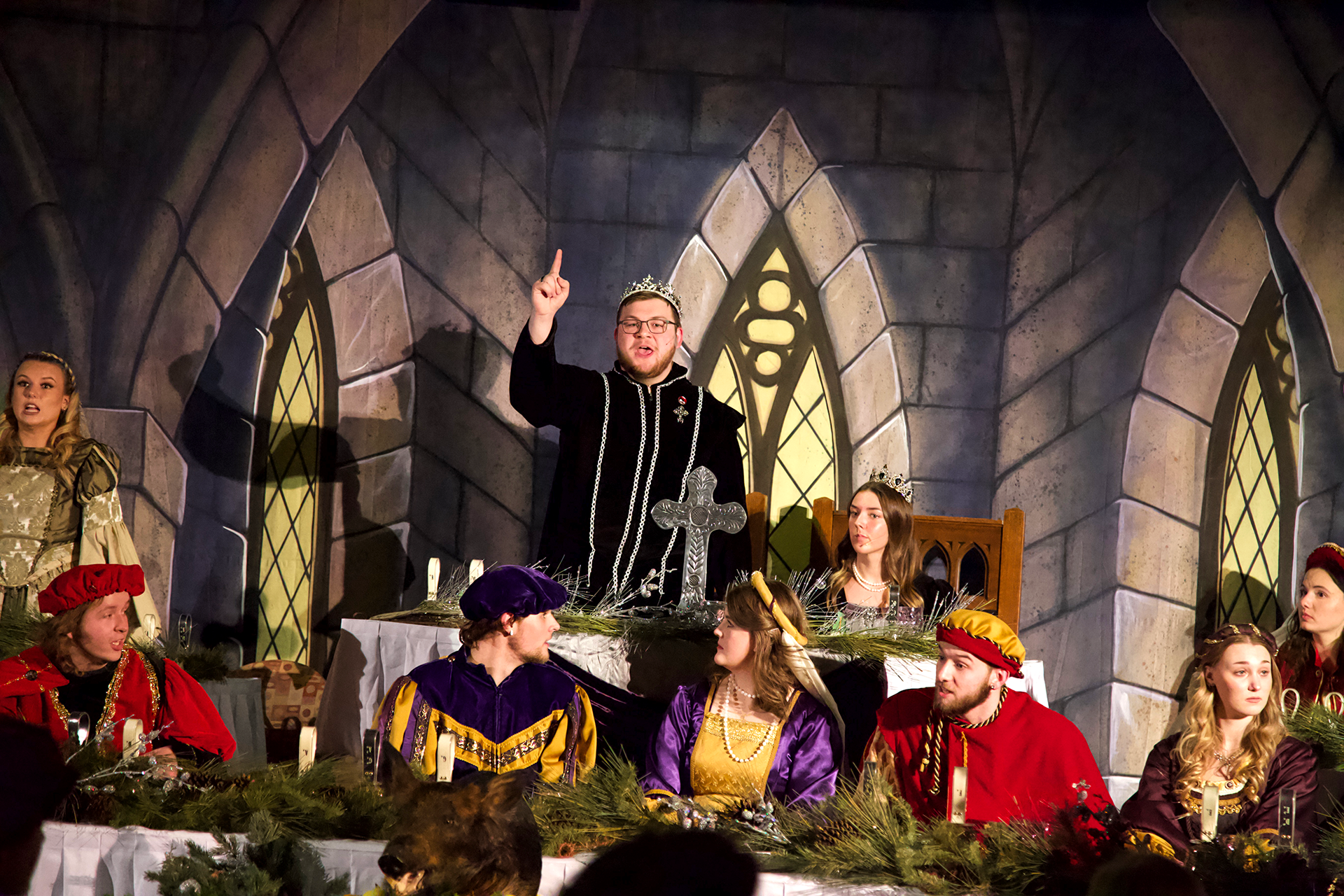 The image shows a medieval-themed banquet with people in elaborate costumes. At the center, a man wearing a crown stands and points upwards, appearing to be giving a speech. Others, dressed as nobles, sit around a long table adorned with evergreen branches.