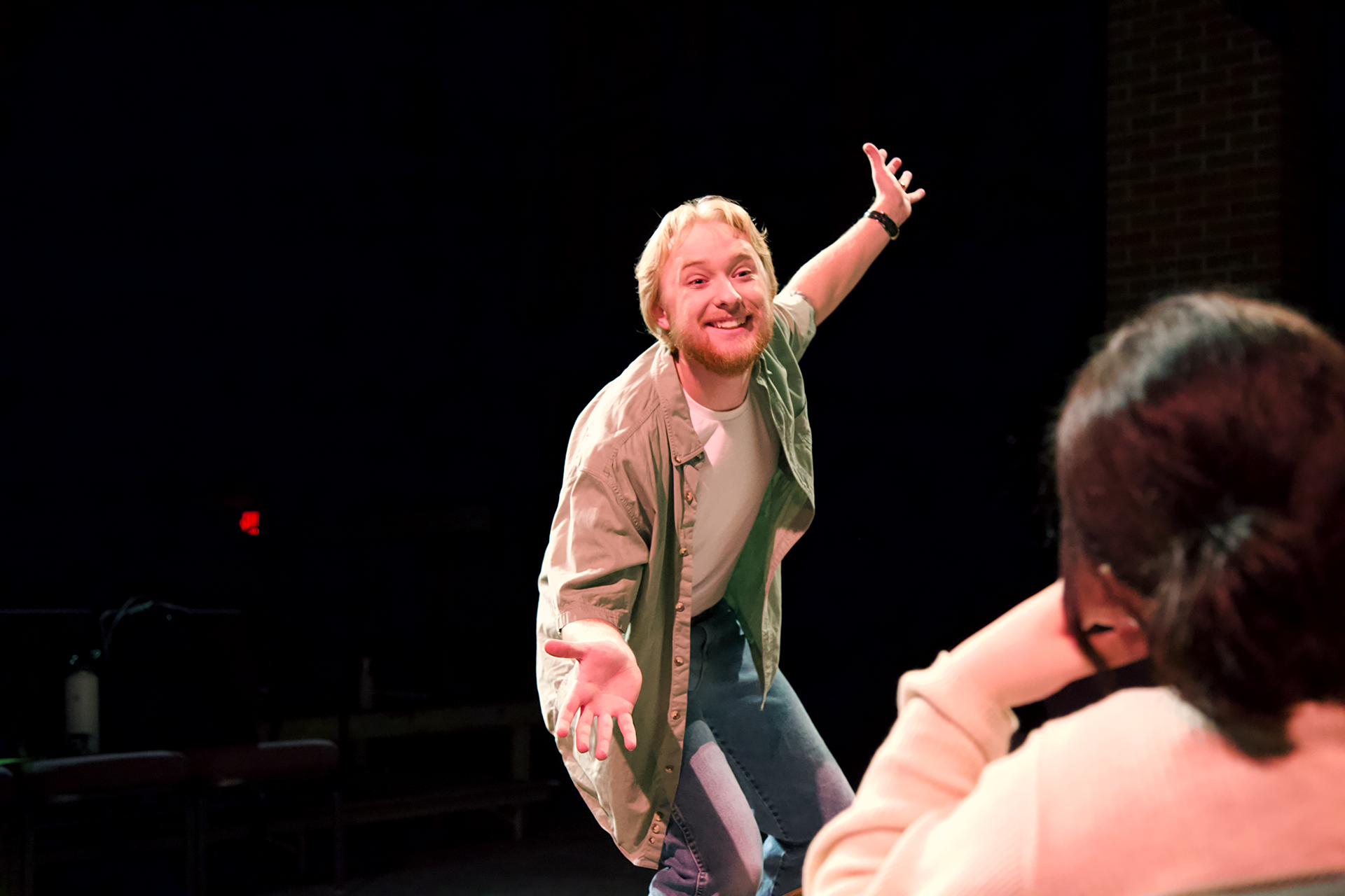 A man with blond hair and a beard is enthusiastically performing on stage, reaching out with both arms wide open. He wears a casual outfit of a beige jacket over a white shirt and blue jeans. A person in the foreground is watching him, resting their chin on their hand.