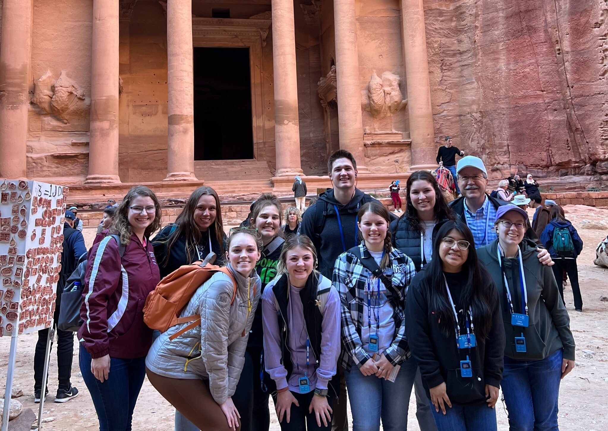 A group of twelve people stands in front of the ancient rock-cut architecture of Petra's Treasury in Jordan. They are smiling and posing for the photo, with some wearing lanyards around their necks. The sandstone facade of the Treasury is detailed and illuminated by sunlight.
