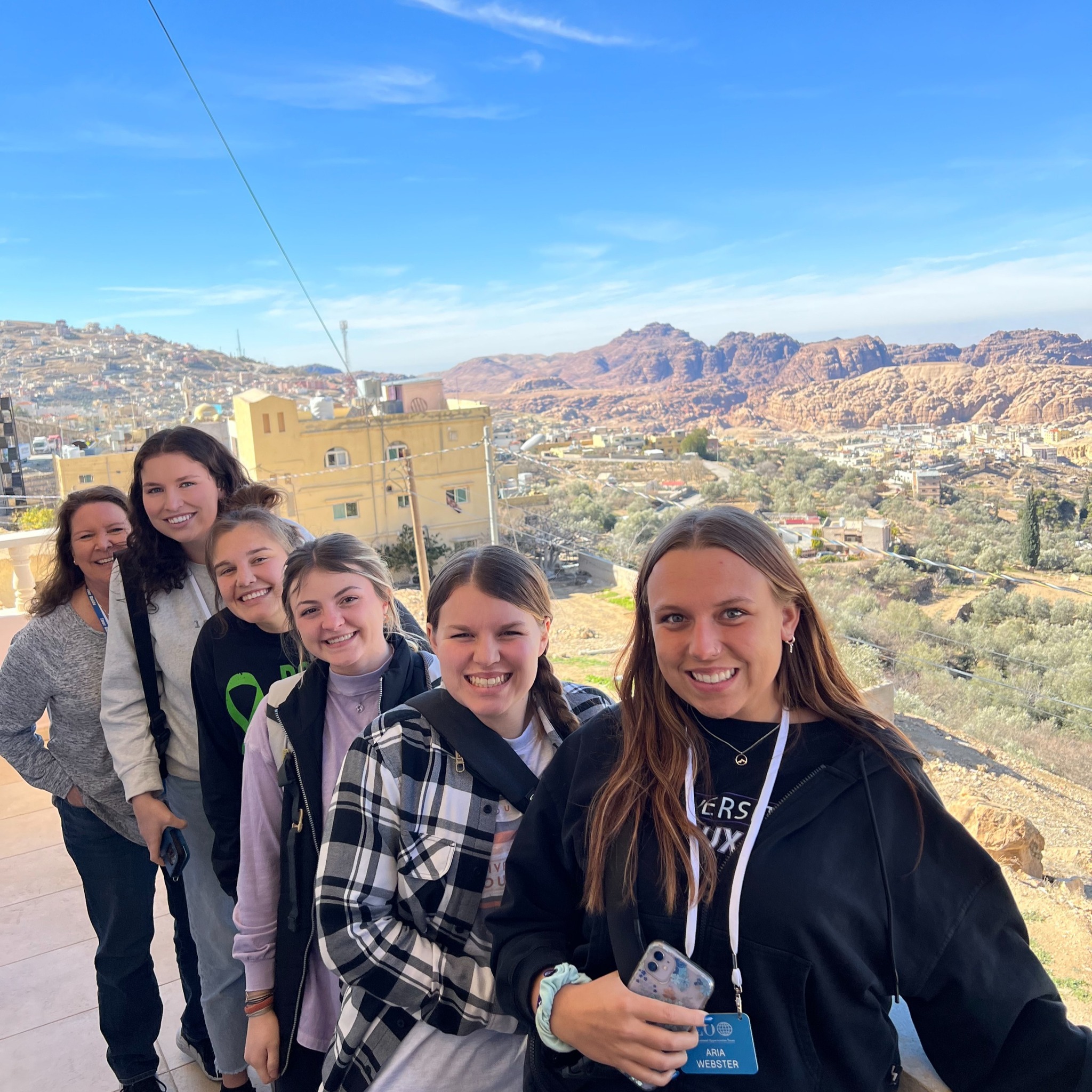 A group of five people, four young women and one older woman, stand closely together on a balcony overlooking a scenic landscape. The view features a mix of buildings, greenery, and distant rocky mountains under a clear blue sky. All of them are smiling towards the camera.