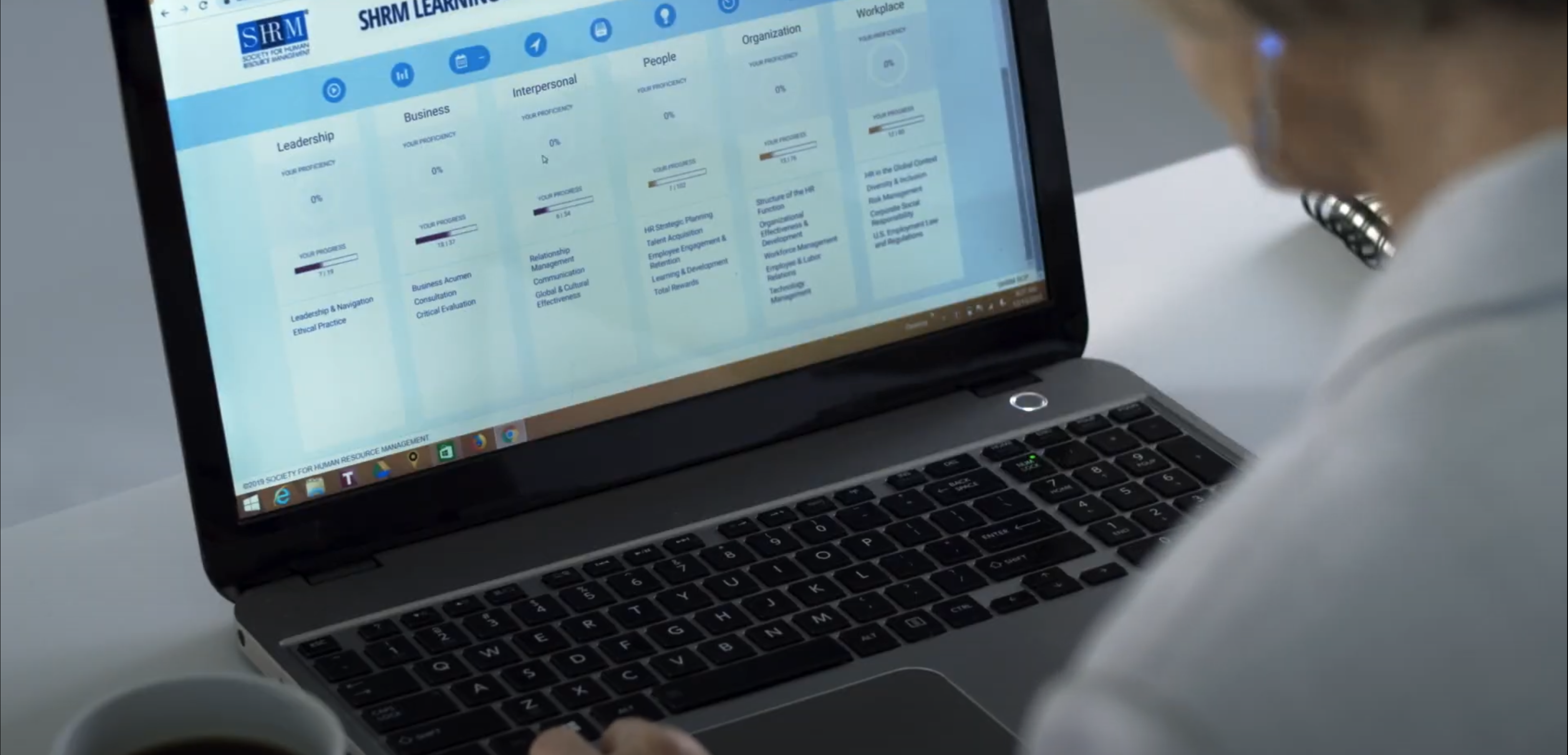 A person is seated at a desk, using a laptop. The screen displays a web page with multiple columns labeled "Leadership," "Business," "Interpersonal," "People," and "Workplace," suggesting an e-learning platform. A cup of coffee is visible on the left side of the desk.