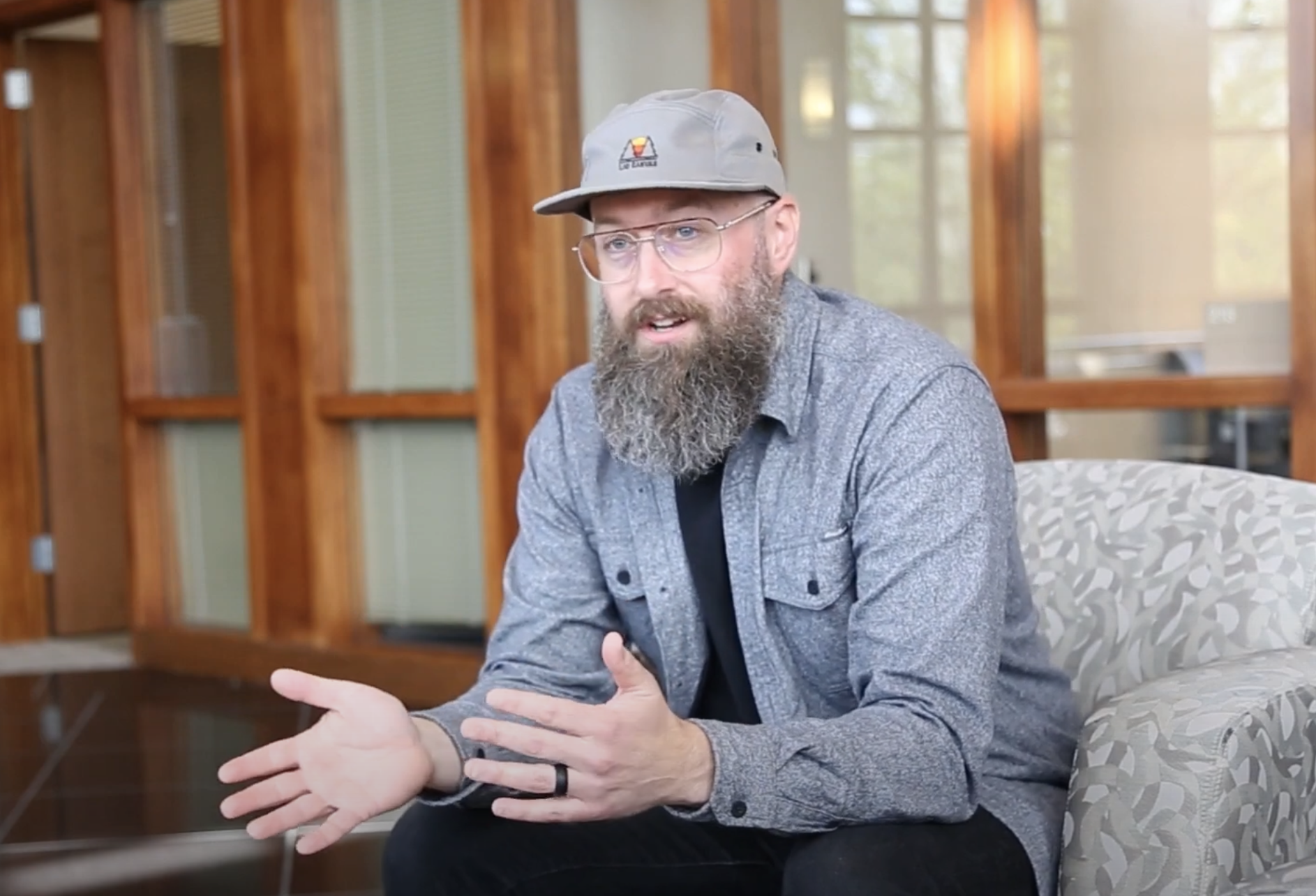 A bearded man wearing glasses and a gray cap, dressed in a gray button-up shirt and black pants, sits on a patterned chair in a modern room with wooden accents. He is gesturing with his hands as he speaks.