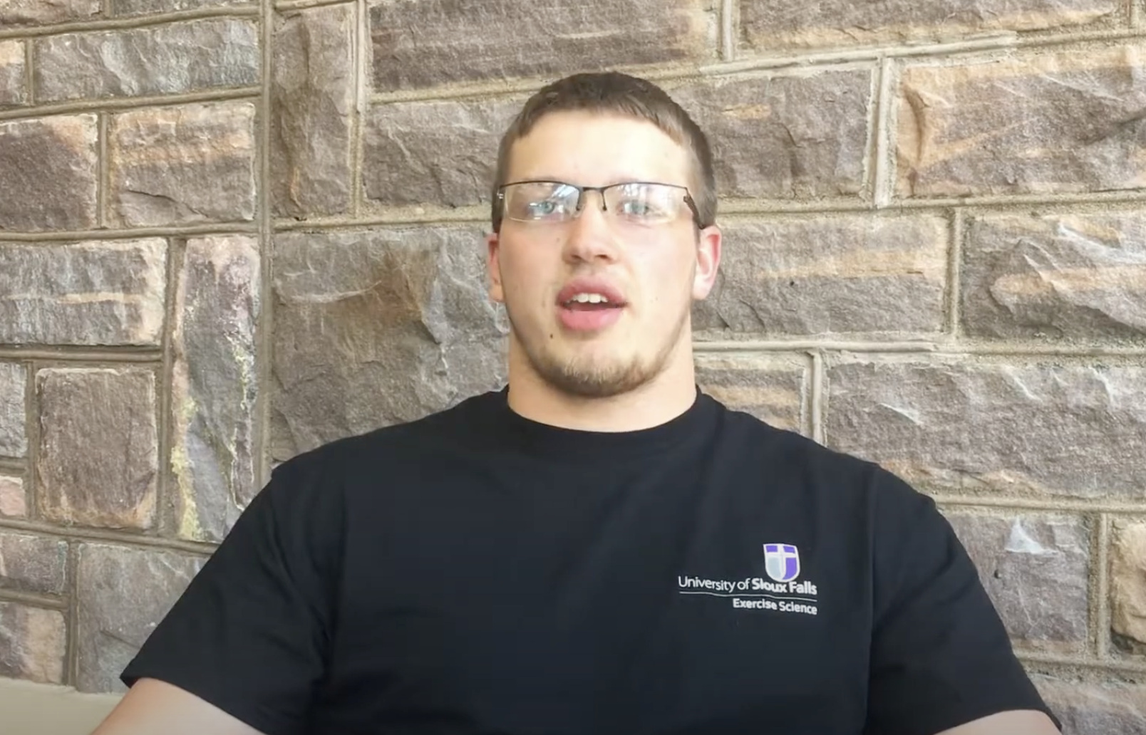 A young man with short hair and glasses wears a black T-shirt with "University of Sioux Falls Exercise Science" written on it. He is sitting against a stone wall. He appears to be talking or explaining something.