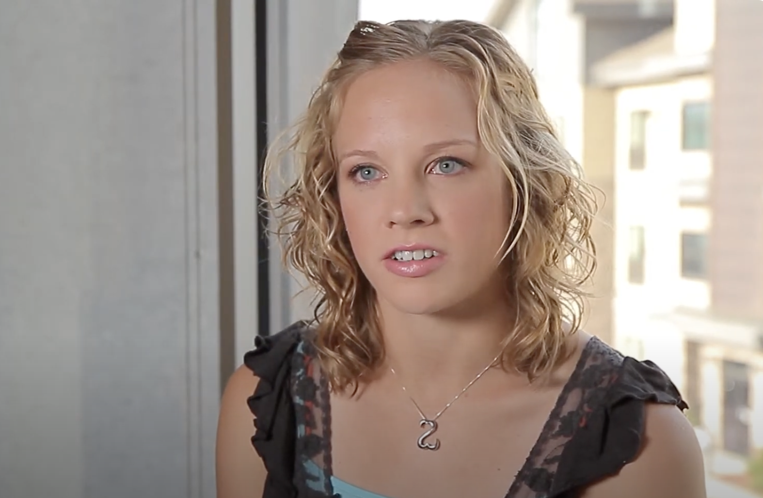 A woman with wavy blonde hair is sitting indoors near a window. She is wearing a black ruffled sleeveless top over a light blue tank top and a silver necklace with a snake pendant. She appears to be in her mid-conversation or explaining something.