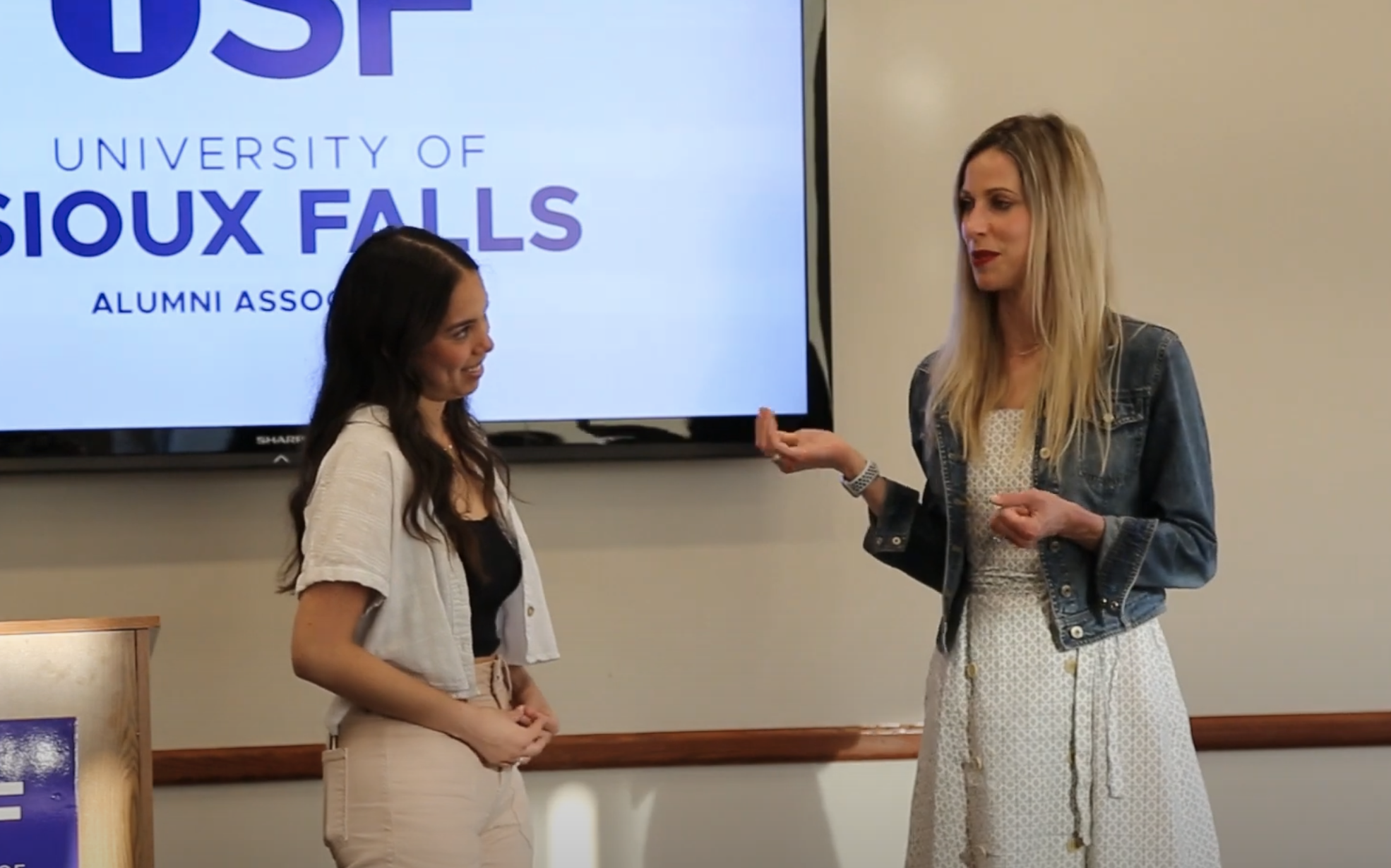 Two women stand in front of a screen that reads "University of Sioux Falls Alumni Association." One woman, wearing a black top and white pants, listens as the other, in a white dress and denim jacket, speaks. They are likely engaged in a discussion or presentation.