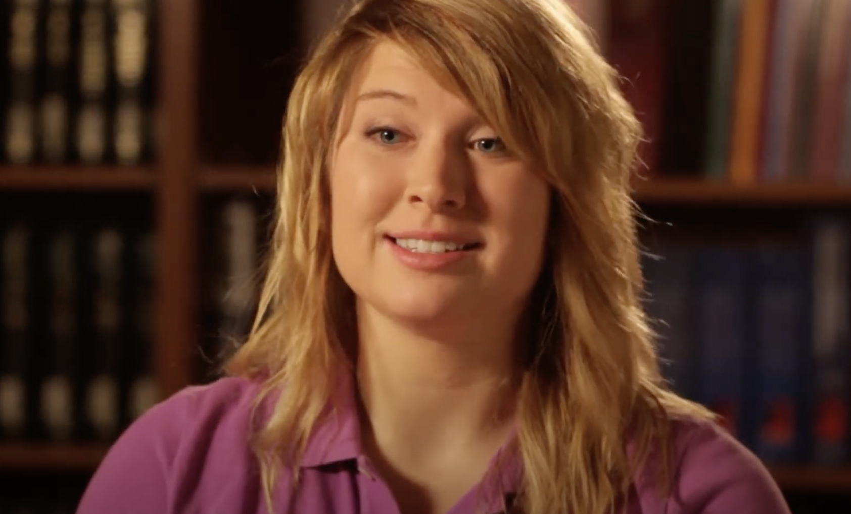 A young woman with long blonde hair smiles at the camera. She is wearing a purple collared shirt and is positioned in front of a bookshelf filled with books. The lighting highlights her face and softens the background.