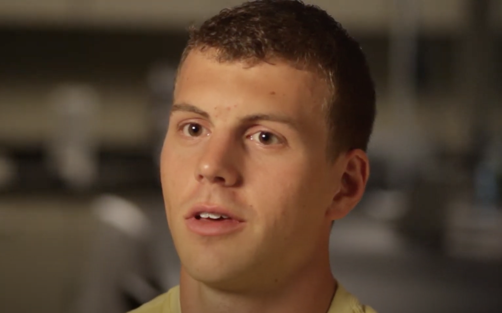 A young man with short brown hair is speaking. He is wearing a light-colored shirt. The background is blurred, focusing the viewer's attention on his face.