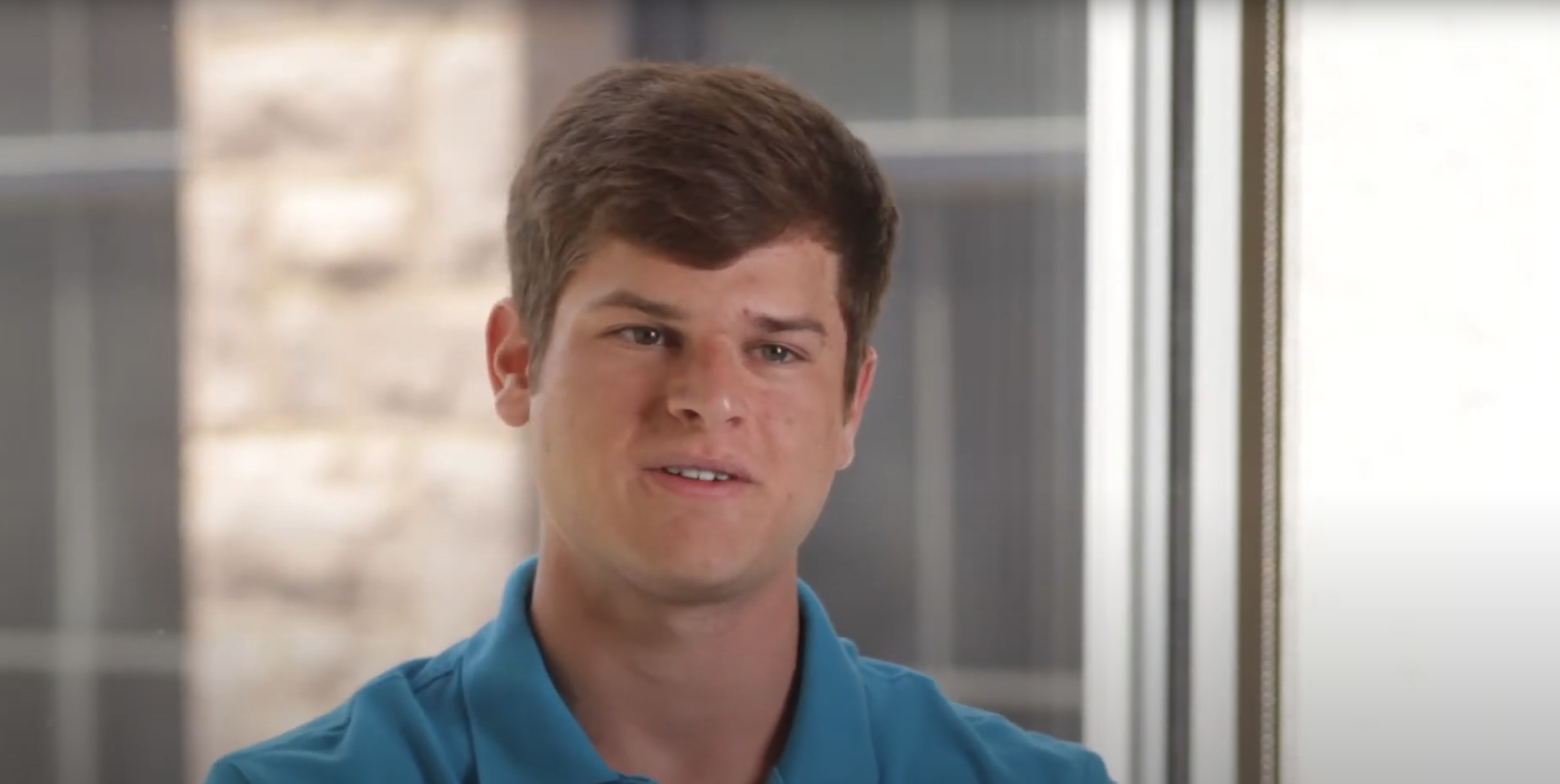 A young man with short brown hair and a blue polo shirt sits indoors by a window, looking slightly to the side. The background features a blurred view of stone wall and window panes.