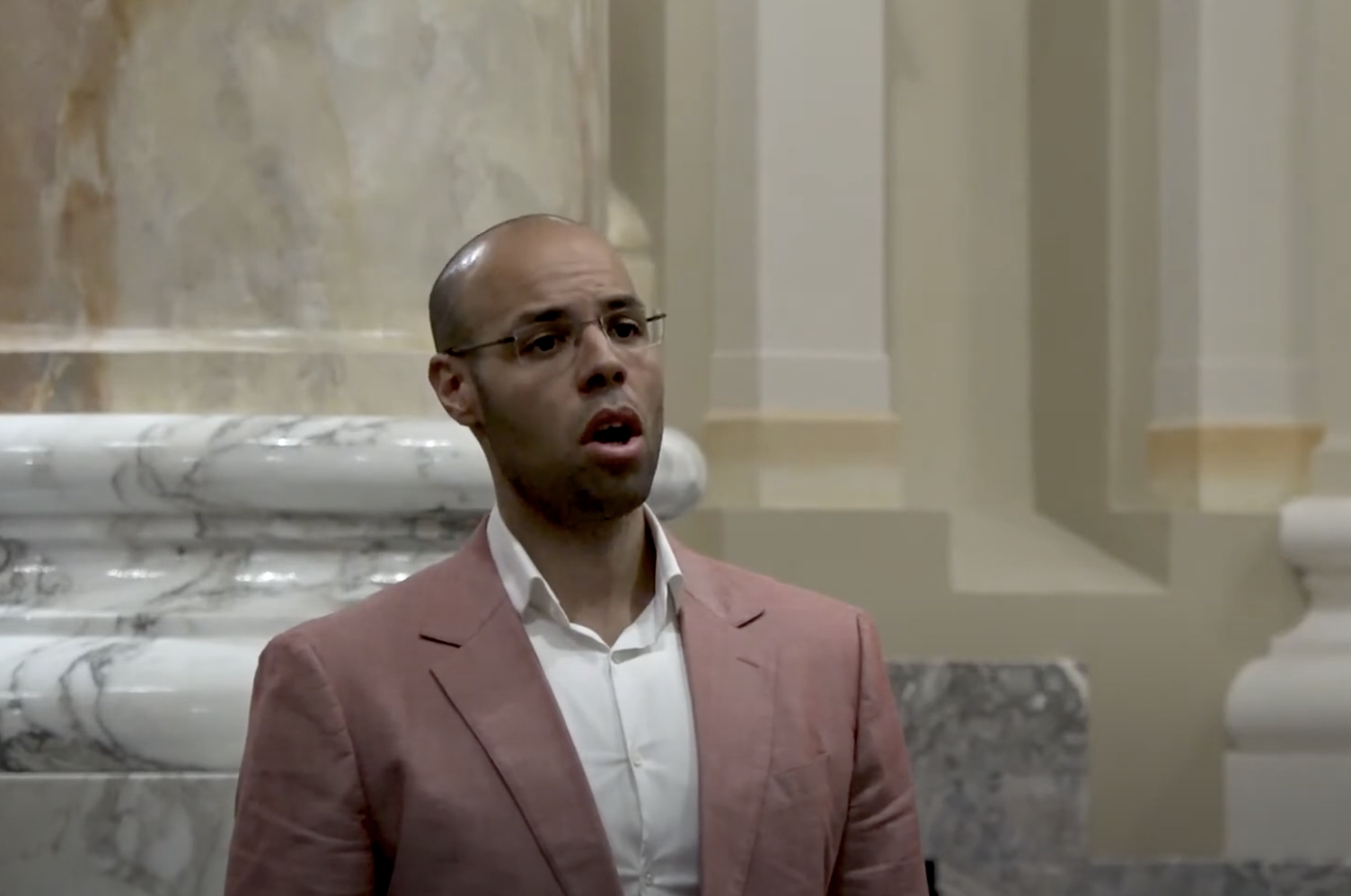 A man with a bald head, glasses, and a beard is seen wearing a pink blazer and a white shirt. He appears to be singing or speaking passionately. The background features ornate marble columns and light-colored walls, suggesting an elegant indoor setting.