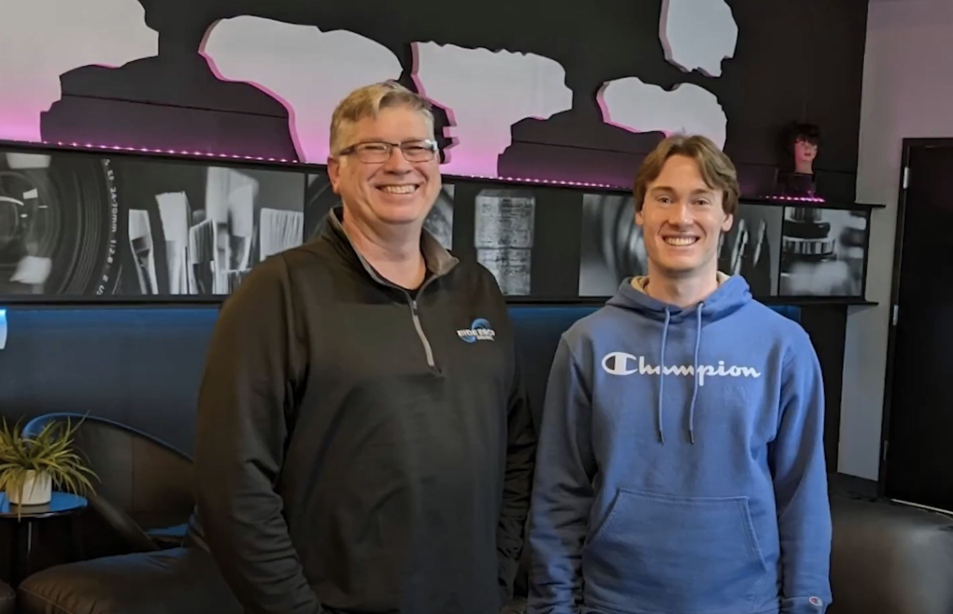 Two people smiling and standing side by side in front of a mural with black, white, and pink colors. The person on the left is wearing glasses and a black sweatshirt, while the person on the right is wearing a blue "Champion" hoodie. The background includes some plants and decor.